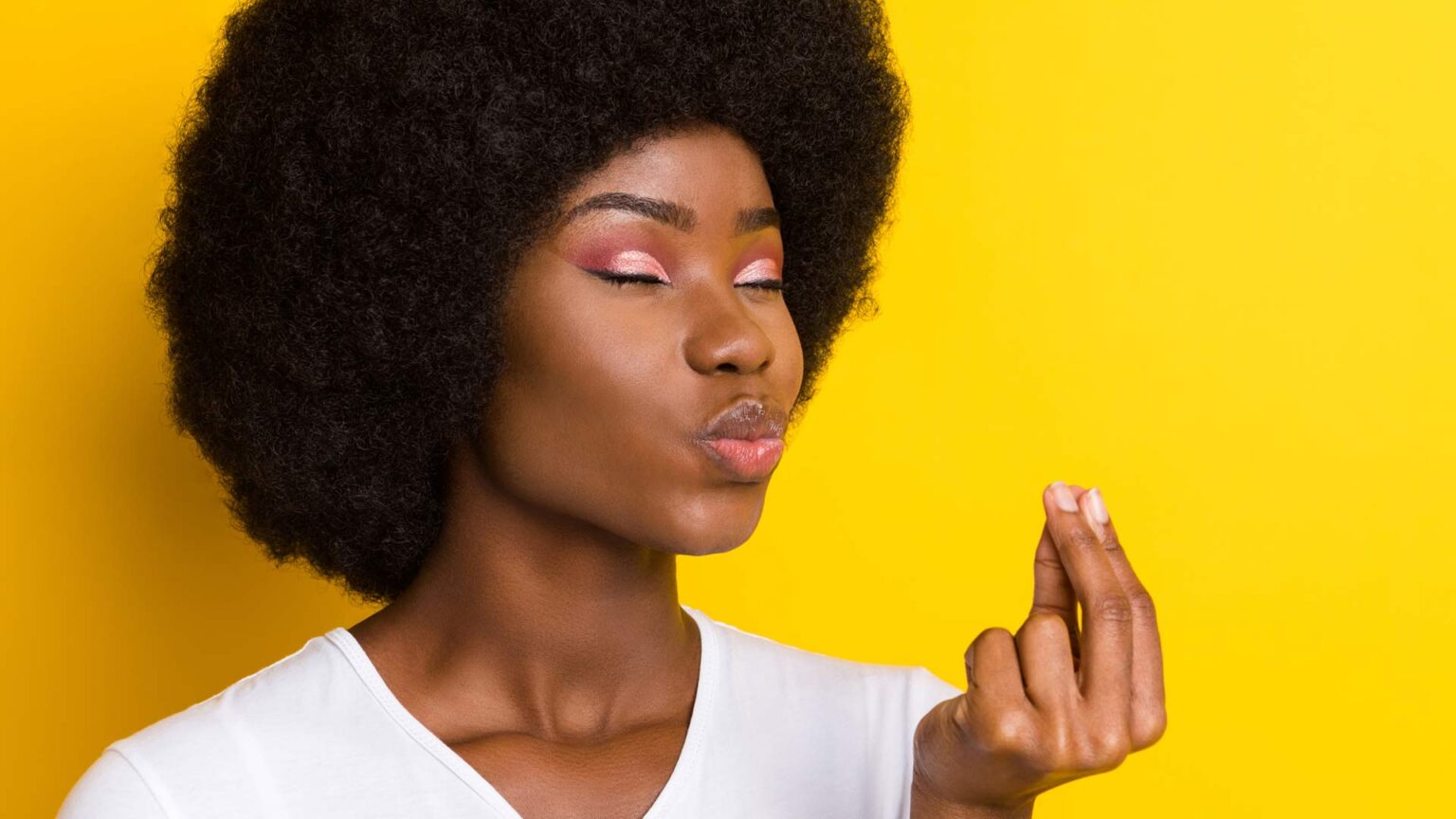 A dark-skinned woman with closed eyes and pursed lips holding her forefingers together against a yellow background.