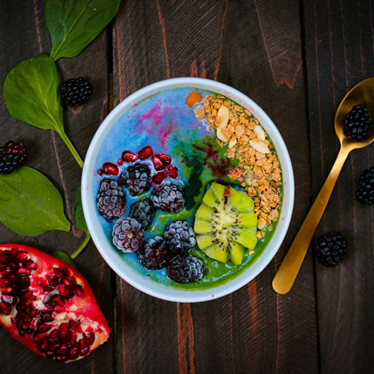 A top-down view of a vibrant smoothie bowl served in a white bowl on a dark wooden table. The bowl is topped with blackberries, pomegranate seeds, kiwi slices, and granola over a colorful blue and green base. A sliced pomegranate and fresh spinach leaves are scattered nearby.