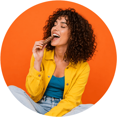 Person with curly hair smiling while enjoying a chocolate snack bar against an orange background.