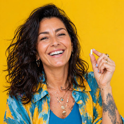 A portrait of a smiling, middle-aged woman with dark curly hair against a bright yellow background. She is holding up a small white supplement capsule between her fingers, representing health supplementation and proactive aging.