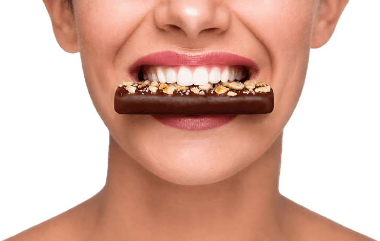 Close-up of a smiling woman biting a chocolate-coated snack bar against a blue background.
