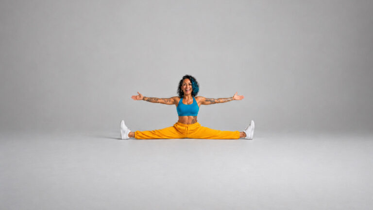 Tattooed woman with blue curly hair doing the splits with arms outstretched and a big smile, wearing a blue sports top and yellow pants against a gray background.