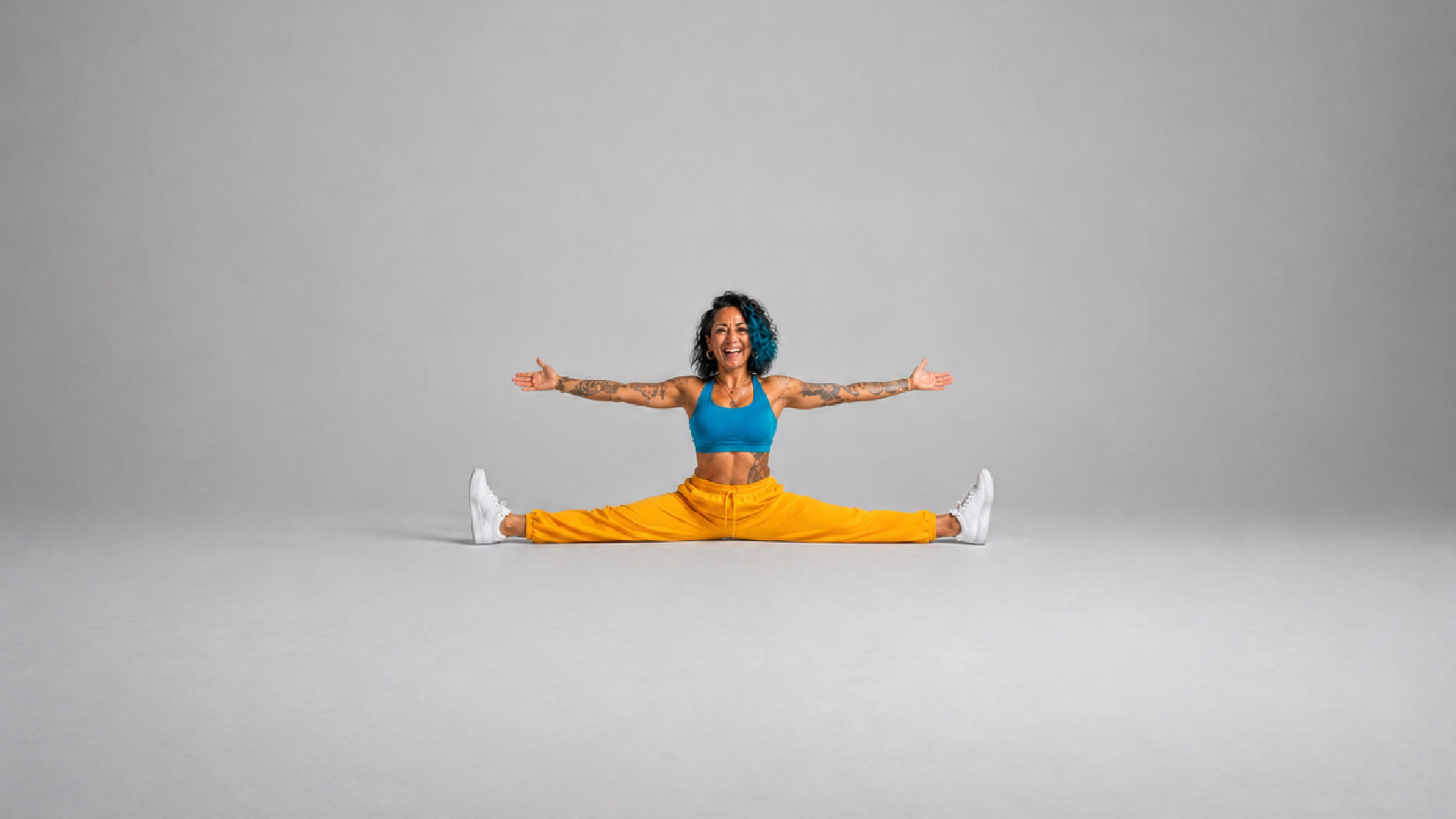Tattooed woman with blue curly hair doing the splits with arms outstretched and a big smile, wearing a blue sports top and yellow pants against a gray background.